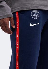 Άνδρας με σκούρο μπλε παντελόνι Paris Saint-Germain της Nike με κόκκινη ρίγα με το λογότυπο της Nike και λευκό λογότυπο Nike swoosh στον μηρό.