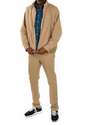 Mann trägt beige Jacke und Hose, blaues Hemd mit Knöpfen und schwarze Turnschuhe, schließt den Reißverschluss der Jacke, steht vor weißem Hintergrund.