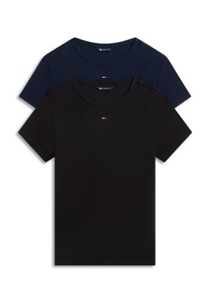 Frau trägt eine dunkel bordeauxrote Jacke, schwarzes Hemd, blaue ausgestellte Jeans und schwarze Schuhe mit quadratischer Spitze, stehend vor einem einfarbigen Hintergrund.