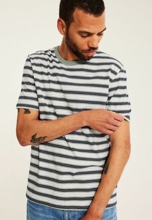 Mand med kort krøllet hår og skæg iført en sort-hvid stribet t-shirt, som justerer sin ærme, synlige blå jeans.