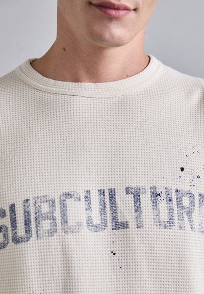 Felpa in maglia waffle di un beige chiaro con stampa del testo "SUBCULTURE" in blu sbiadito, con delicate spruzzate di dettagli blu.