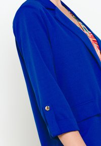 Modrý blazer s hladkou texturou, s ohrnutými rukávy a zlatým knoflíkem. Obsahuje klopový límec a přední kapsy.