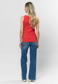 Červený žebrovaný top bez rukávů s kulatým výstřihem, doplněný o světle modré široké džíny. Pozadí je jednobarevné bílé, zaměřující se na detaily oblečení.