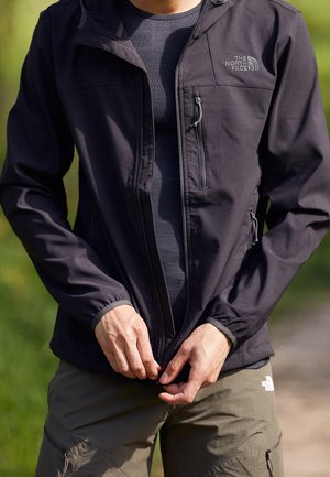 Personne portant une veste foncée et un pantalon olive, fermant la veste à l'extérieur avec un fond vert flou.
