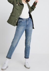 Chaqueta de estilo militar verde con varios bolsillos, top blanco ajustado, jeans de tiro alto azul claro y zapatillas blancas con suelas grises.