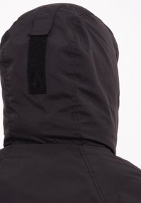 Zwarte hoodiejacket met een matte afwerking, gestructureerde stof en een klittenbandpatch op de achterkant. Naaddetails zichtbaar langs de randen.