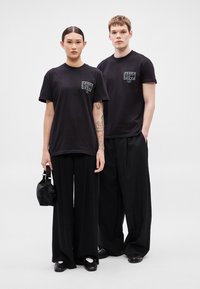 Czarne bawełniane koszulki z krótkim rękawem, z grafiką na kieszeni z przodu. Modele noszą luźne czarne spodnie i dopasowują je do czarnego obuwia.