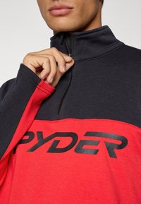 Rood en zwart half-zip pullover sweatshirt met een logo, voorzien van lange mouwen en een gladde textuur. De rits bevindt zich in het midden bij de hals.