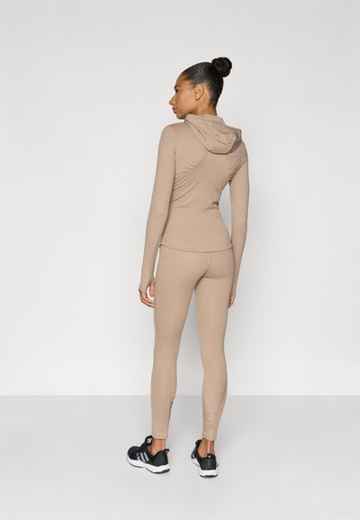 Σετ αθλητικών ρούχων σε μπεζ με εφαρμοστή γραμμή, που περιλαμβάνει μπλούζα με κουκούλα και ταιριαστό κολάν, με σχεδιασμό χωρίς ραφές και ελαφρύ ύφασμα. Μαύρα αθλητικά παπούτσια.
