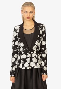 Schwarzer Blazer mit floralem Muster, der über weiße Spitze-Details, schmalen Revers, einen einzigen Knopfverschluss und eine taillierte Silhouette verfügt. Getragen über einem schwarzen Kleid.