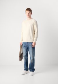 Kremfarget strikket genser med ribbestrikkede mansjetter, blå rette jeans og hvite joggesko. Brun crossover-veske med lang stropp.