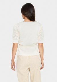 Krēmkrāsas tops ar īsām, teksturētām adītām piedurknēm. Augšdaļa ir ar gludu apdari un elastīgu jostasvietu, kas nodrošina pieguļošu izskatu.