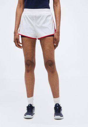 Isik, kellel on seljas valged spordipüksid punase äärisega, tumesinised tossud ja valged sokid, seisab ühevärvilise tausta ees.