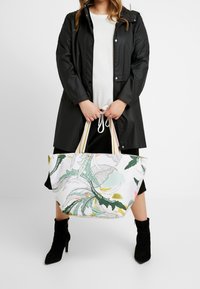 Grand sac cabas en tissu léger avec un motif de feuilles tropicales en vert, jaune et rose. Les poignées rayées apportent une touche colorée.