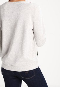 Sudadera gris claro con escote redondeado y mangas largas, que presenta una sutil textura y un detalle bordado en el dobladillo. Combinada con jeans oscuros.