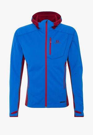 Veste softshell bleue avec fermetures éclair rouges, capuche doublée rouge, poches poitrine et latérales zippées, et étiquette "Windstopper" en bas à l'avant.
