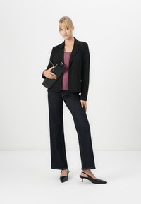 Kvinne iført svart blazer, lilla topp, mørke jeans og svarte hæler, holding en svart veske mot en hvit bakgrunn.