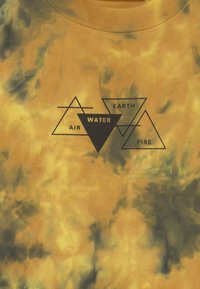 Žluté tričko s efektom tie-dye vyrobené z bavlny, zdobené černými geometrickými potisky: "VZDUCH," "VODA," "ZEM," a "OHEŇ" ve tvaru trojúhelníků.