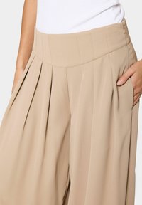 Pantaloni beige a pieghe con vita alta ampia, caratterizzati da linee pulite, una consistenza liscia e una zip laterale invisibile.
