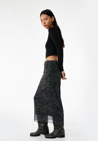 Zwarte lange mouwen crop top gecombineerd met een doorschijnende, gedessineerde rok met donkere groen-tijgerstrepen, gecombineerd met zwarte enkellaarsjes.
