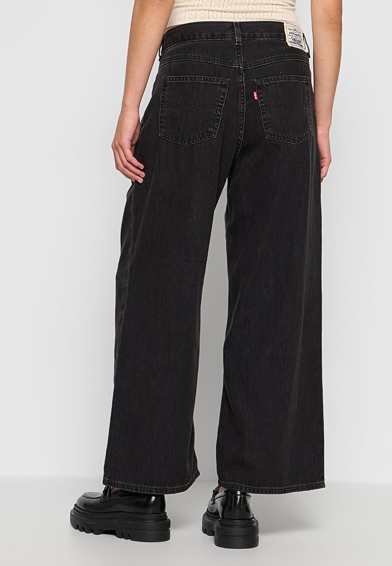 Jean noir à coupe large en denim, avec une taille haute, deux poches arrière et une étiquette logo sur la taille. Texture lisse.