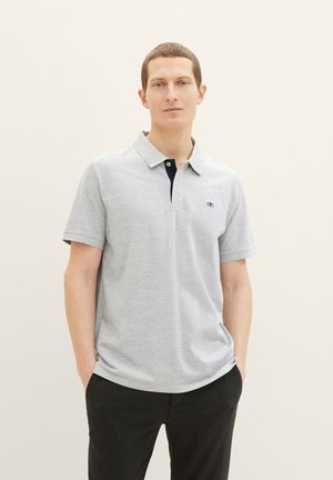 Mężczyzna z krótkimi włosami stoi z rękami w kieszeniach, ubrany w jasnoszarą koszulkę polo i ciemne spodnie na prostym beżowym tle.