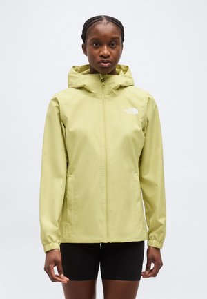 Naine, kes kannab helerohelist kapuutsiga vihmajakki ees lukuga ja musti lühikesi pükse, seisab tavalise heleda tausta ees.