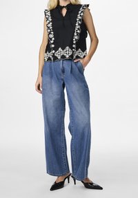 Haut noir sans manches avec broderie florale blanche et bords à volants, associé à un pantalon en denim bleu à jambes larges et des escarpins noirs à talons aiguilles.