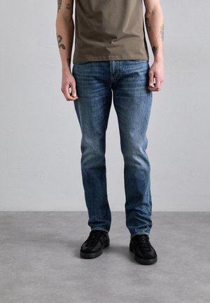 Mand iført blå jeans, sorte sneakers og en brun t-shirt, stående på en grå gulv mod en grå væg.