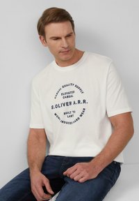 Witte katoenen t-shirt met een ronde afbeelding met donkerblauwe tekst, korte mouwen en een ronde halslijn, gedragen met donkerblauwe spijkerbroeken.