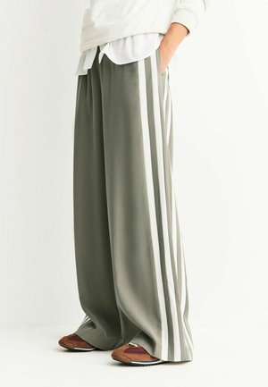 Weite Hosen aus olivgrünem Stoff mit weißen Seitenstreifen. Verfügen über Seitentaschen und werden mit mehrfarbigen Turnschuhen getragen.