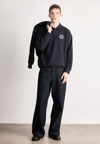 Μπλε ναυτικό πόλο πουλόβερ με κυκλικό λογότυπο, συνδυασμένο με φαρδιά σκουρόχρωμα τζιν και μαύρα παπούτσια, κρατώντας μπουφάν στον έναν ώμο.