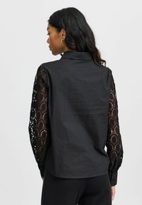 Schwarzes Shirt mit solidem Rücken und kunstvollen langen Ärmeln aus Spitze mit einem floralen Muster. Verfügt über einen Kragen und geknöpfte Manschetten.