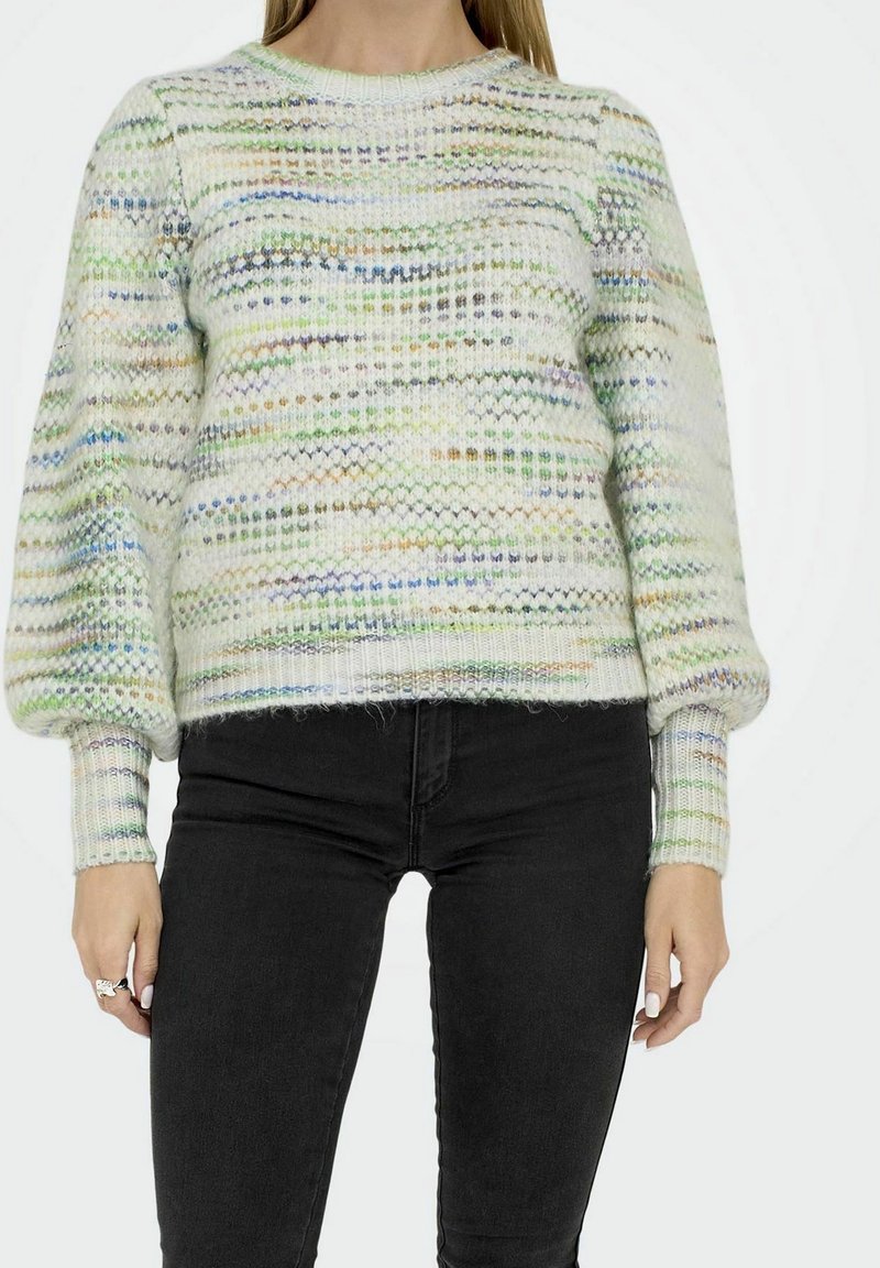 Femme portant un pull tricoté multicolore et texturé avec des manches ballon et un pantalon noir ajusté, sur un fond uni.