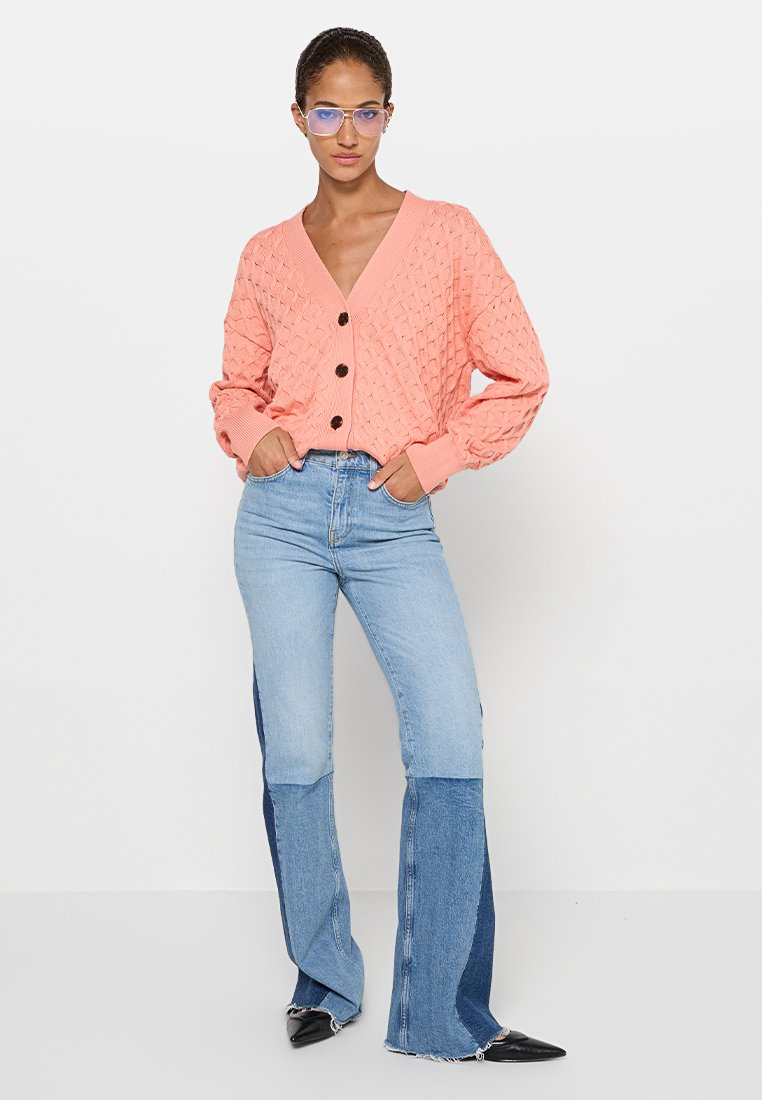 Miękki różowy sweter wykonany na drutach z wzorem w diamenty, posiadający dekolt w kształcie litery V i czarne guziki, połączony z dzwonowymi niebieskimi dżinsami z blokowaniem kolorów.