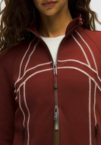 Giacca zip-up color bordeaux con dettagli in cucitura bianca, colletto alto e due tasche laterali con zip. Tessuto morbido e liscio con un design aderente.