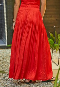 Jupe maxi plissée rouge avec une taille élastique, texture lisse et design fluide. Portée avec des sandales noires sur une surface en gravier.