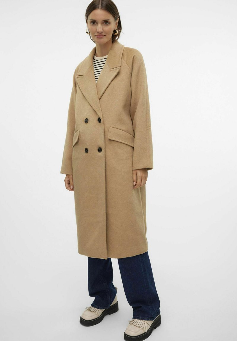 Manteau long en mélange de laine beige avec un devant croisé, des revers à pointe, des boutons noirs et des poches latérales, porté sur un top rayé.