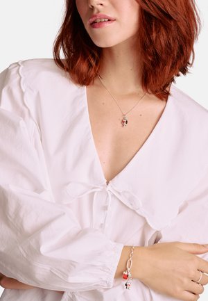 Silberne Halskette mit einem bunten Anhänger, getragen zu einer weißen Bluse mit gewelltem Ausschnitt und Puffärmeln.