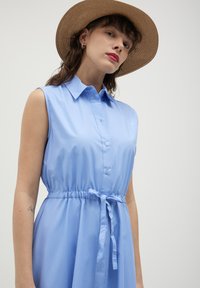 Abito senza maniche azzurro chiaro con colletto, chiusura a bottoni davanti e cintura elastica con dettaglio annodato. Tessuto morbido con una leggera lucentezza.