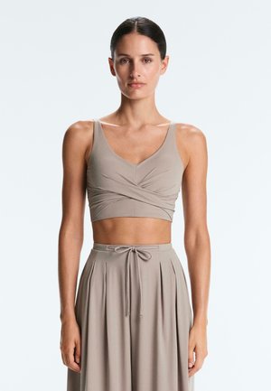 Sötéthajú nő bézs színű, átkötős stílusú crop topban és magas derekú, redőzött szoknyában, amelynek övvel kötött derékrésze van, egyszínű háttér előtt áll.