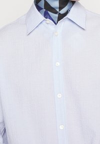 Světle modrá texturovaná košile s knoflíkovým zapínáním vpředu a bílými knoflíky, doplněná o vzorovaný tmavě modrý šátek na krku.