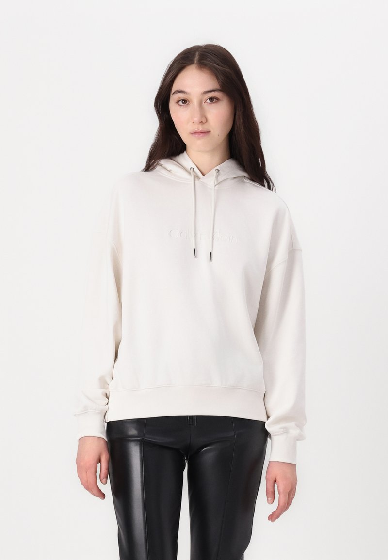 Felpa beige con vestibilità rilassata, maniche lunghe e cappuccio con coulisse. Presenta un logo goffrato sottile sul petto e polsini a coste.