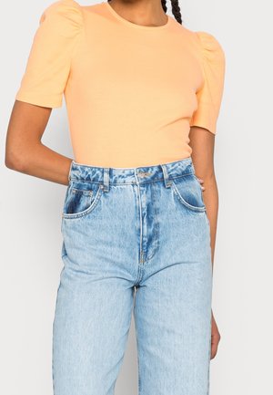 Persoon draagt een lichtoranje top met pofmouwen en een lichtblauwe high-waisted spijkerbroek, staat met een arm gebogen achter de rug.