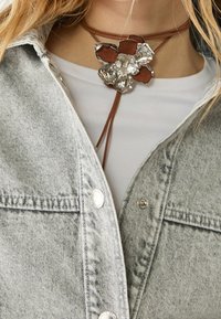 Hnědý kožený náhrdelník s kovovým přívěskem ve tvaru květiny, který má texturované okvětní lístky a hnědý akcent, nošený přes šedou džínovou bundu.