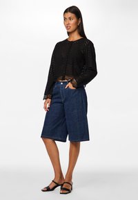 Cardigan nero all'uncinetto con maniche lunghe, abbinato a shorts in denim blu a metà coscia. La modella indossa sandali neri con tacco.