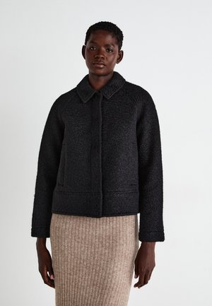 Veste noire texturée coupée avec un col, fermeture à boutons sur le devant et deux poches latérales, assortie à une jupe beige côtelée.