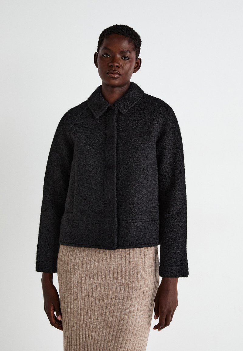 Black, textured cropped jacket with a collar, front button closure, and two side pockets, paired with a ribbed beige skirt.