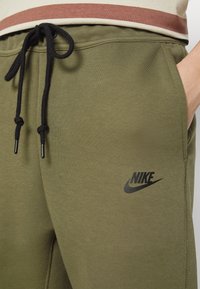Olivu zaļas Nike sporta bikses, kas izgatavotas no mīksta auduma, ar melnu logo uz augšstilba un melnām auklām, lai pielāgotu derību.