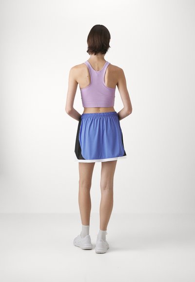 Lilla atletisk crop top kombinert med en blå skort, med svarte sideaksenter. Modellen står med en nøytral bakgrunn, iført hvite sko.